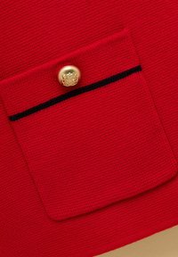 Lähikuva punaisesta neulotusta kangastaskusta, jossa on musta reunus ja kultainen nappi, jossa on kohokuvioitu vaakunakuva.