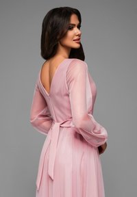 Lång rosa klänning med långa, genomskinliga ärmar och djup V-back. Har ett knytmidd som betonar midjan och flödande tyg för en graciös siluett.