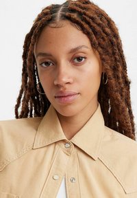 Camicia di velluto a coste beige con colletto appuntito, dotata di bottoni a scatto e dettagli cuciti. Indossata con una maglietta sotto, che mette in mostra il tessuto testurizzato.