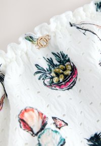 Tissu en œillet blanc à bord froufroutant, imprimé de coquillages colorés et d'un panier de plantes, avec un petit fermoir circulaire doré.