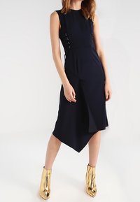 Marinblå ärmlös klänning med asymmetrisk fåll, sidoknytning, slät tygtextur, ihop med blanka guldankelstövlar.