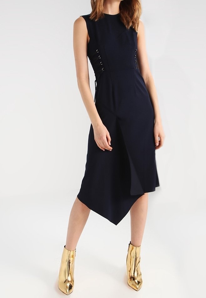 Marinblå ärmlös klänning med asymmetrisk fåll, sidoknytning, slät tygtextur, ihop med blanka guldankelstövlar.