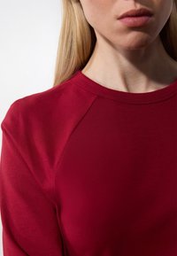 Piros, hosszú ujjú felső, kerek nyakkivágással, sima anyagból készült. Rágland ujjakkal rendelkezik, kiemeli a testhez simuló dizájnt és a tiszta varrást.