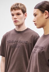 To personer iført brune bomulds-T-shirts med broderet tekst "TOMMY JEANS". Enkel rund halsudskæring, afslappet pasform, minimalistisk design.
