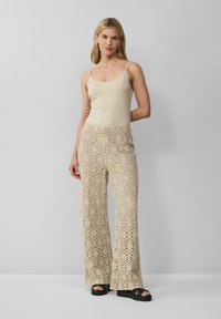 Beige tanktop met dunne bandjes, gecombineerd met wijdvallende haakwerk broeken met een hexagonaal patroon. Zwarte platform sandalen maken de look af.