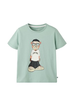 T-shirt vert clair avec un chien dessin animé portant des lunettes de soleil, une casquette noire, une chemise blanche avec le texte "Cool Boy", un pantalon large et des baskets.