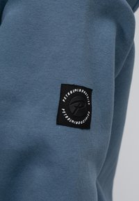 Tessuto di felpa blu con una consistenza morbida. Presenta un'etichetta quadrata nera con testo bianco e logo sul braccio sinistro.