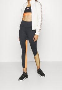 Chaqueta blanca con cremallera y banda con logo, top corto negro con banda naranja, leggings a bloques de color negro y naranja, y zapatillas deportivas negras.