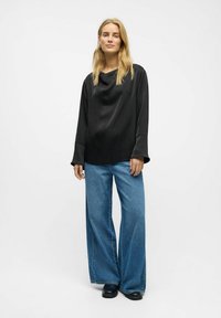 Schwarze Satinbluse mit weitem Ausschnitt und langen, ausladenden Ärmeln, kombiniert mit hellblauen, weiten Jeans und schwarzem Schuhwerk.