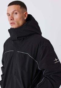 Man met kort donker haar, gekleed in een zwart jasje met een hoge kraag en reflecterende biezen, kijkt lichtjes opzij.