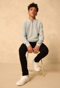 Light blue zippered sweater with a textured fabric, paired with black slim-fit pants and white sneakers, seated on a stool against a beige backdrop.