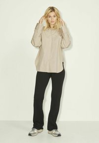 Camicia beige con bottoni, maniche lunghe e tasca, abbinata a pantaloni neri e sneakers grigie. La camicia presenta una vestibilità comoda e una texture liscia.