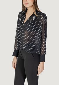 Blusa nera trasparente con pois, caratterizzata da una pattina, texture plissettata e polsini arrotondati. Abbinamento mostrato con pantaloni neri aderenti.