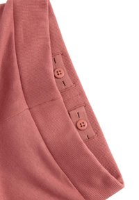 Kankaan vaaleanpunaisessa ribbitekstuuri, jossa on kaksi nappia kiinnitettynä helman tukevaan nauhaan. Muotoilu on pehmeä ja jämäkkä, ja siinä on hienovarainen tekstuuri.
