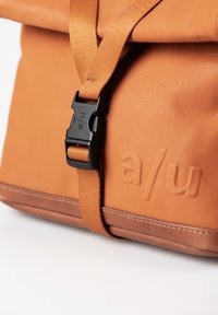 Zaino in tela arancione con una fibbia in plastica nera, con branding impresso e bordo marrone a contrasto lungo la base.