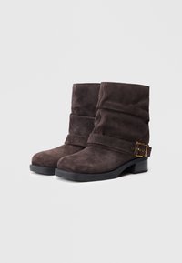 Botas de tobillo de ante marrón con un diseño relajado, que cuentan con una correa ancha y detalles de hebilla en tono dorado, sobre un bajo tacón negro.