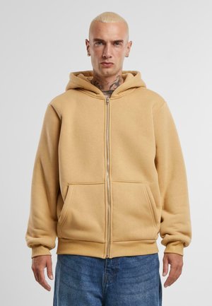 Bež zip-up dukserica od mekanog materijala, sa prednjim zatvaračem, dva prednja džepa i kapuljačom s podešavanjem. Ležeran, opušten kroj.