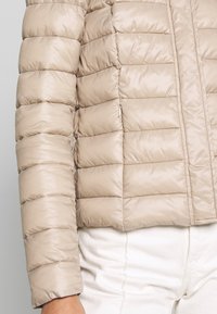 Giacca puffer beige con texture a trapunta e segmenti orizzontali. Presenta un colletto alto e una chiusura con zip sul fronte.