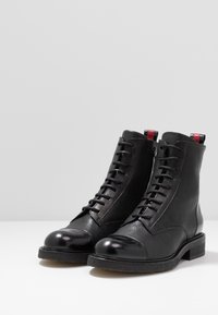 Botas de tobillo de cuero negro con un acabado texturizado, diseño con cordones, punta redondeada de charol y acentos rojos y negros en el tacón.