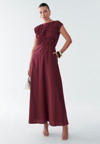 Maxi šaty v tmavom bordovom odtieni, s nariaseným živôtikom, krátkymi rukávmi, vreckami a voľným strihom. Doplněné jednoduchými šperkami.