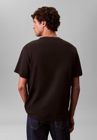 Tumepruun lühikeste varrukatega t-särk ümara kaelusega, valmistatud pehmest kangast. Seljaosa on sile ja mahukas, ilma nähtavate logodeta.