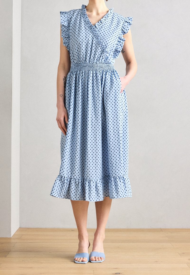 Femme portant une robe midi bleu clair à pois noirs, manches et ourlet à volants, taille cintrée, et talons ouverts bleus, debout sur un sol en bois.