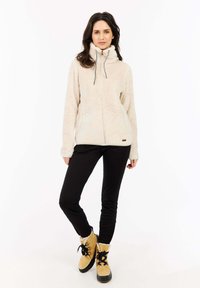 Fleece ritsjack in lichtbeige met hoge kraag, trekkoorden en voorzakken, gecombineerd met zwarte broek en gele laarzen.