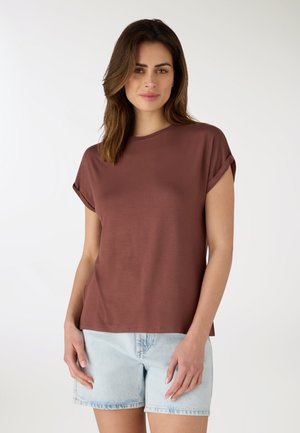 Donna con capelli castani alla lunghezza delle spalle, indossa una maglietta marrone a maniche corte e pantaloncini di jeans azzurri, in piedi davanti a uno sfondo bianco uniforme.