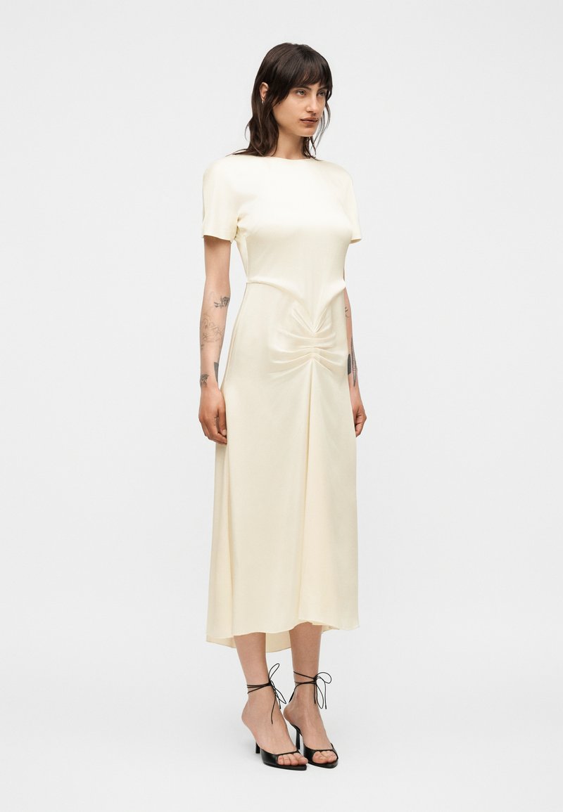 Femme aux cheveux foncés porte une robe midi crème à manches courtes, fronces sur le devant et talons noirs à brides sur un fond blanc uni.