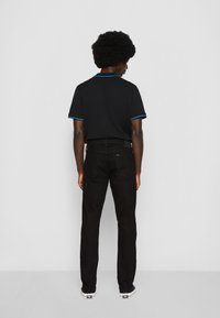 Afro soenguga omav mustad teksad ja must polo-särk sinise äärisega, seistes silmitsi lihtsa valge seina.