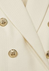 Krémová textúrovaná tkanina na sako s dvojradovými zlatými gombíkmi, každý s podrobným emblémom leva a koruny.