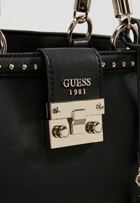 Sac à main en cuir noir avec un fermoir doré, des clous décoratifs et un logo. Comprend des accents métalliques et une finition texturée.