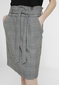 Jupe grise à carreaux taille haute avec une ceinture nouée et des poches latérales, portée avec des mains posées de manière décontractée à l'intérieur des poches.