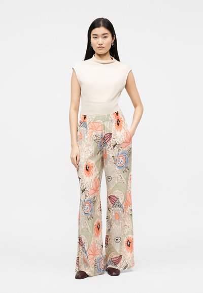 Γυναίκα που φοράει αμάνικο κρεμ τοπ με ψηλό λαιμό και φαρδιά παντελόνια με πολύχρωμο λουλουδένιο μοτίβο, στέκεται με το ένα χέρι στην τσέπη.