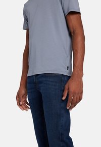T-shirt grigio a maniche corte realizzato in tessuto morbido, abbinato a jeans blu scuro. Il T-shirt presenta un'etichetta a dettaglio di branding nel bordino.