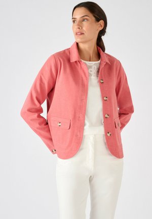 Femme portant une veste corail boutonnée sur un haut en dentelle blanc et un pantalon blanc, se tenant avec les mains derrière le dos devant un fond uni.