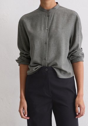 Personne portant une chemise grise boutonnée aux manches retroussées et rentrée dans un pantalon noir, sur un fond clair uni.