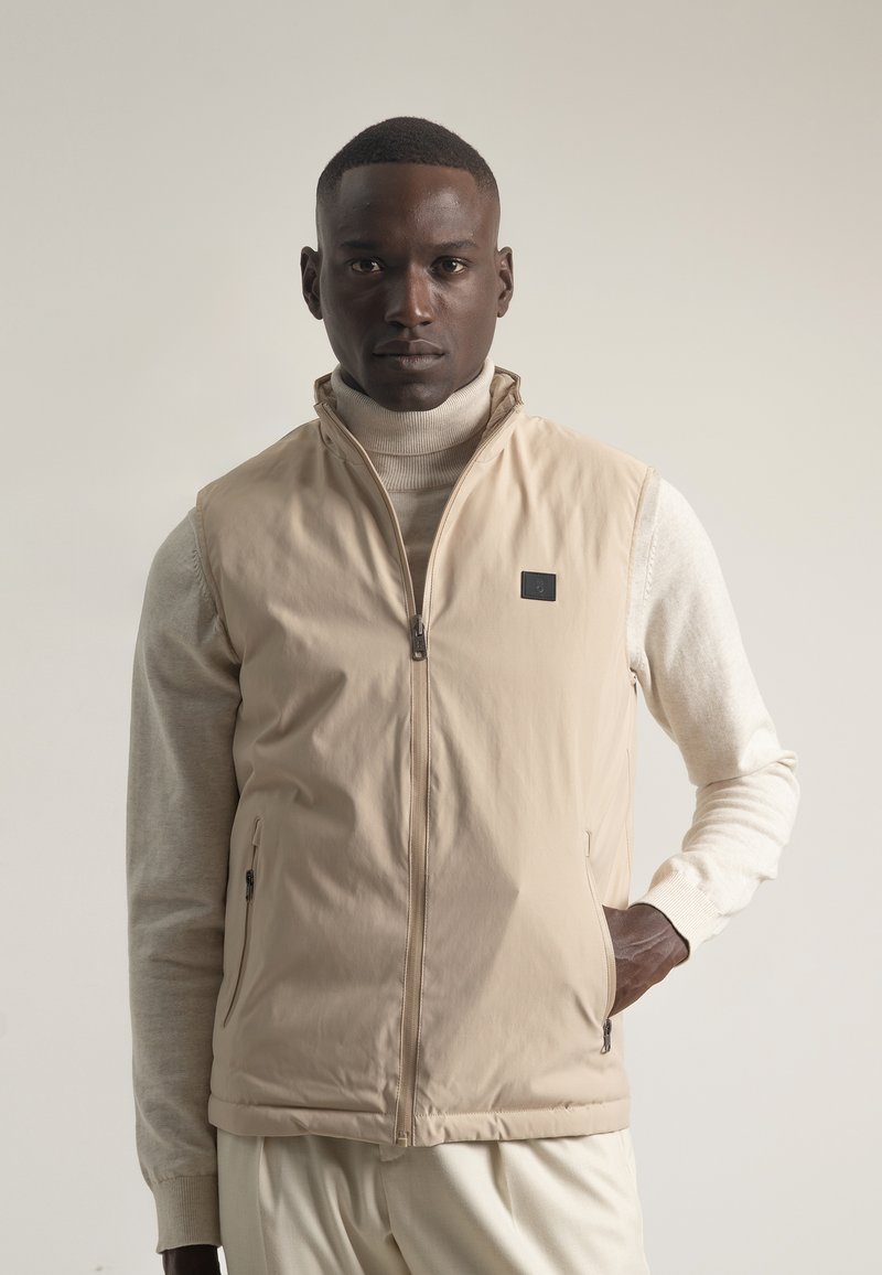 Chaleco beige con cuello alto y cierre frontal, hecho de material suave. Cuenta con bolsillos laterales y una etiqueta de marca discreta.