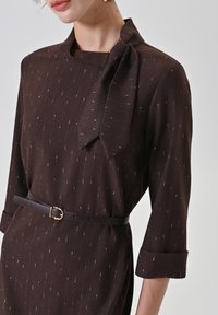 Robe en tissu marron avec un col montant, ornée d'un nœud et de fines rayures verticales. Complétée par une ceinture fine en cuir et des manches retroussées.