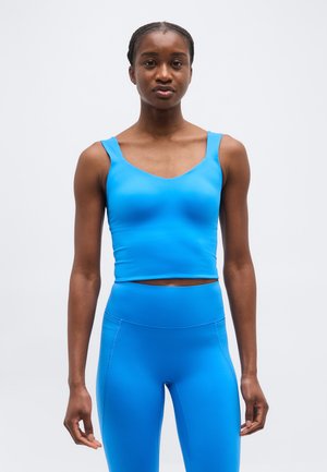 Vrouw staand, gekleed in een felblauwe, mouwloze cropped top en bijpassende, hoge taille leggings tegen een effen lichte achtergrond.