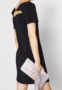 Vestido negro de manga corta con un corte en la espalda, combinado con un clutch plateado con lentejuelas que presenta un detalle anudado y flecos.