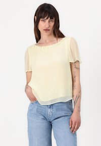 Blouse jaune à manches courtes et transparente avec un accent floral au niveau du col. Associée à un jean taille haute bleu clair.