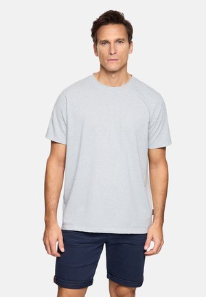 Hombre con cabello corto y castaño, vestido con una camiseta de manga corta gris claro y pantalones cortos azul oscuro remangados, de pie frente a un fondo liso.