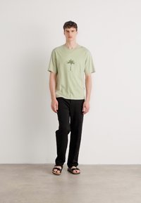 Lys grøn t-shirt med et palme træ grafik, parret med sorte bukser og brune sandaler, stående mod en ensfarvet baggrund.