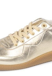 Zapatilla metálica dorada con textura suave, puntera redonda y cierre de cordones beige; presenta un logo y una suela de goma con patrones de tracción.