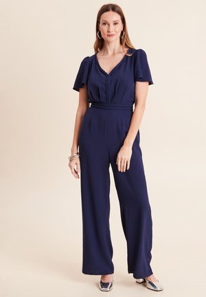 Frau trägt einen marineblauen Overall mit kurzen Ärmeln, V-Ausschnitt, geflochtenem Taillenband, silbernen Blockabsatzschuhen und geometrischen Ohrringen vor einem beigen Hintergrund.