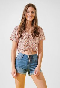 Pamukasti crop top s malim ružičastim i ljubičastim cvjetnim uzorkom, V-izrezom i dekorativnom vezicom na struku, u kombinaciji s svijetloplavim trapericama.