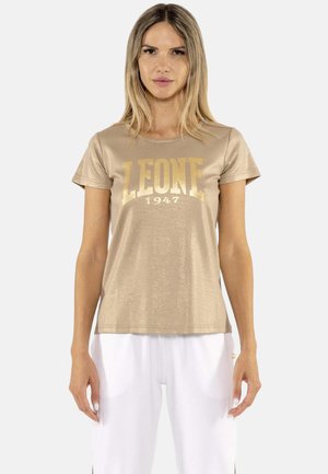 Donna con capelli biondi lisci indossa una t-shirt metallizzata dorata "LEONE 1947" e pantaloni bianchi, in piedi davanti a uno sfondo chiaro e uniforme.