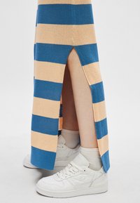 Jambes en chaussettes blanches et baskets visibles à travers la fente latérale d'une jupe tricotée rayée bleu et beige sur fond blanc.