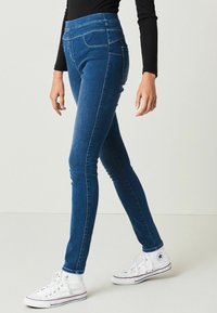 Džínové skinny jeans v tmavě modré barvě s vysokým pasem, se dvěma kapsami a hladkou texturou, spárované s bílými vysokými teniskami.